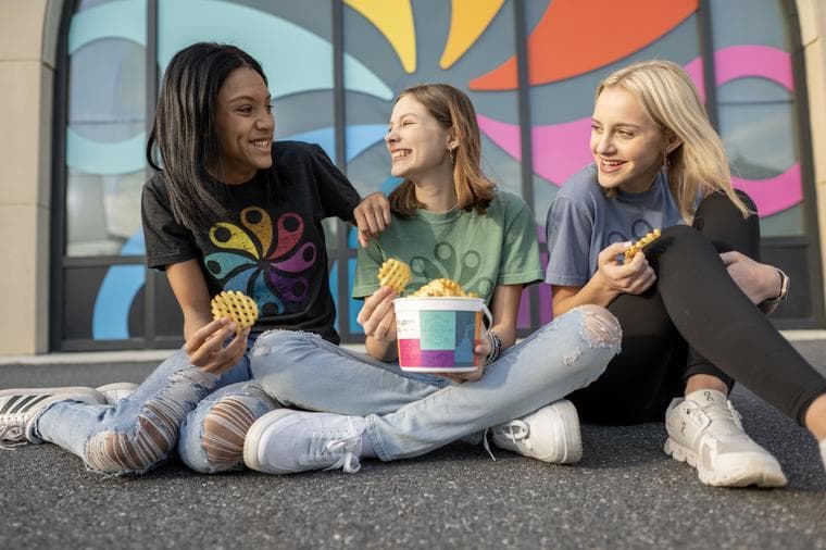 3 girls sitting on the pavement in front of a pinwheel logo, eating french fries